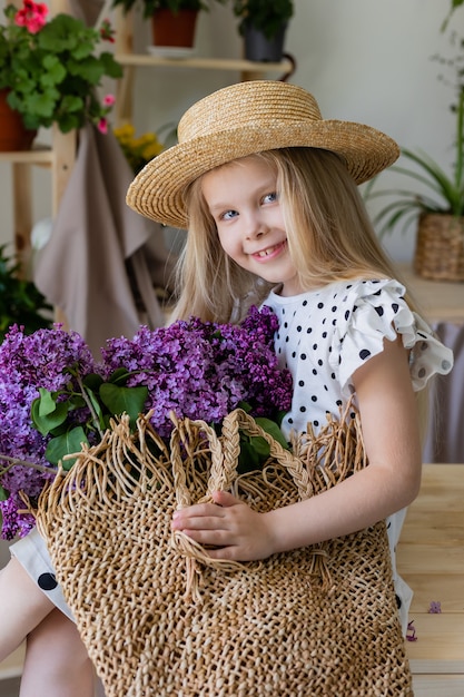 Portrait d'un bébé blond dans un chapeau de paille avec un bouquet de lilas dans un panier en osier dans ses mains