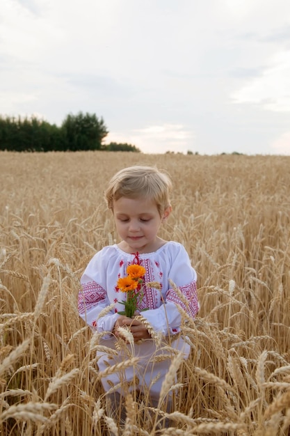 Portrait de bébé en chemise avec broderie rouge et bouquet de fleurs à la main dans un champ de blé au coucher du soleil