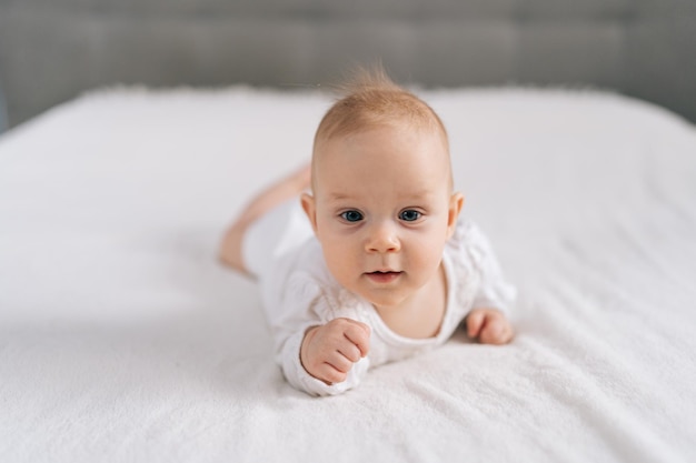 Portrait d'un bébé curieux et doux dans un maillot blanc allongé sur le ventre agite joyeusement les bras et