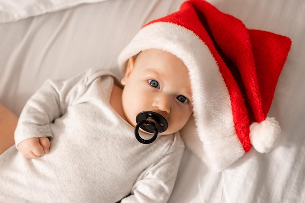 Portrait d'un bébé dans un body blanc et bonnet de Noel avec une tétine noire dans sa bouche allongée sur un oreiller blanc. charmant enfant aux yeux bleus. espace pour le texte. Photo de haute qualité