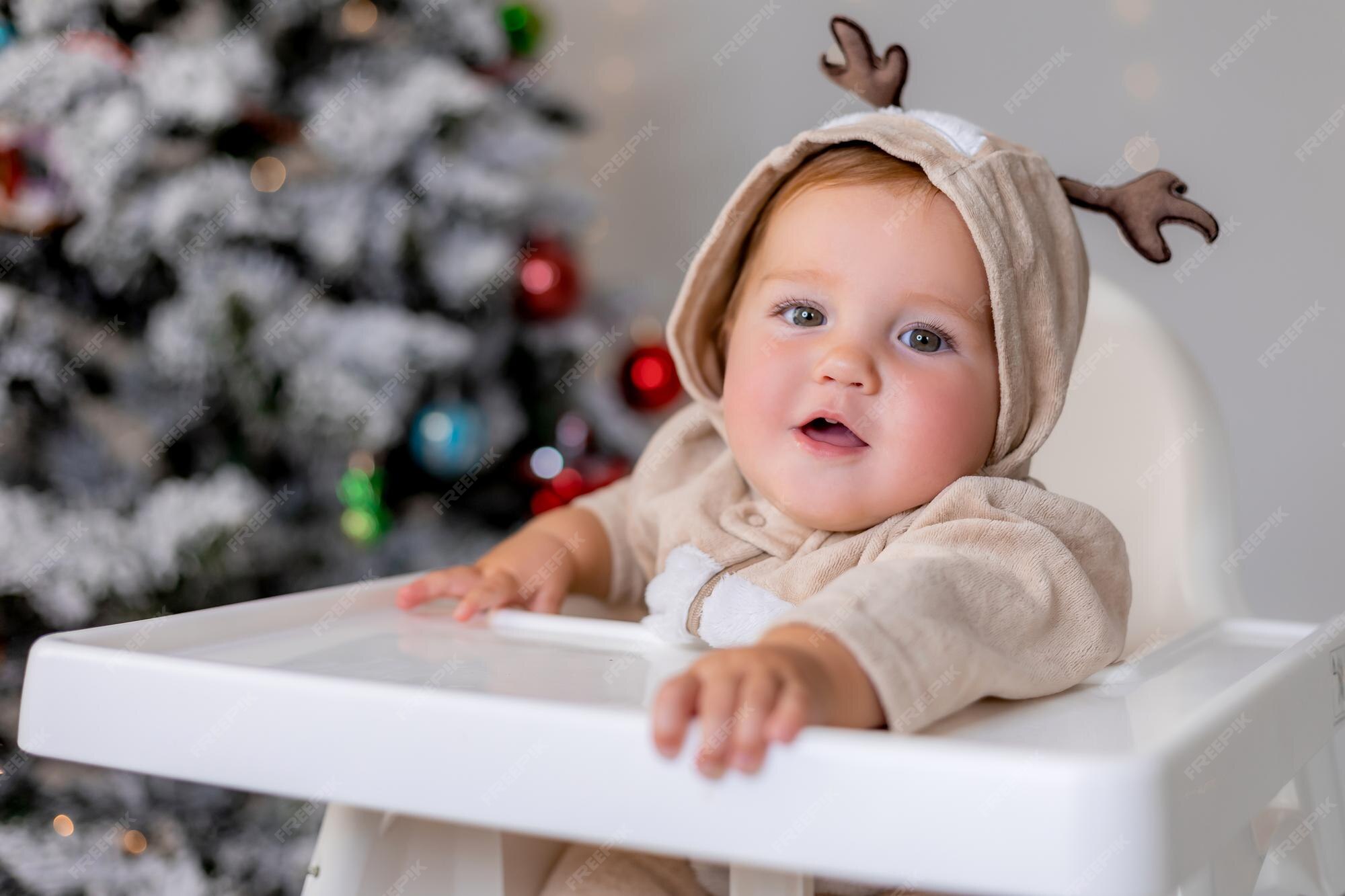 Portrait D Un Bebe Joufflu A Joues Dans Une Combinaison Avec Des Cornes De Cerf Est Assis Dans Une Chaise Haute Blanche Pour Se Nourrir Sur Le Fond De L Arbre De Noel Concept
