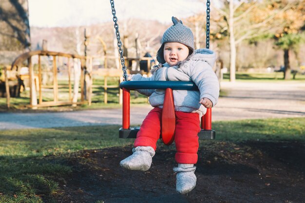 Portrait d'un bébé mignon vêtu de vêtements chauds sur une balançoire à l'aire de jeux par une journée ensoleillée à l'extérieur.