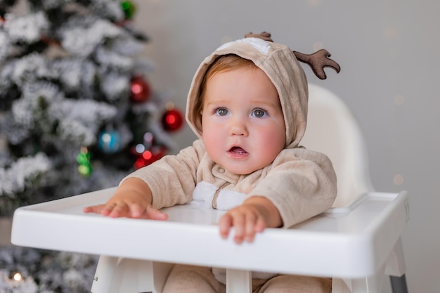 portrait de bébé potelé dans une combinaison avec des cornes de cerf est assis dans une chaise haute blanche près de l'arbre de Noël