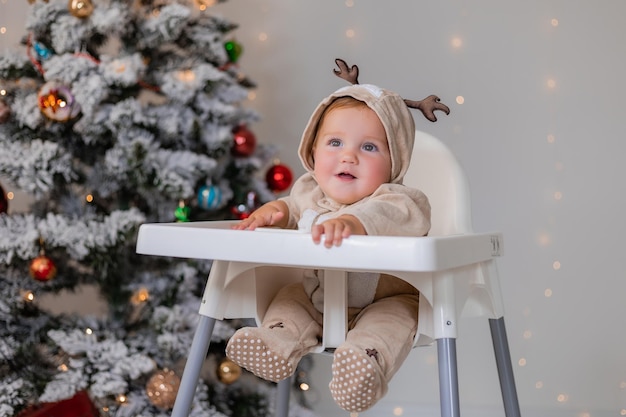 portrait de bébé potelé dans une combinaison avec des cornes de cerf est assis dans une chaise haute blanche près de l'arbre de Noël