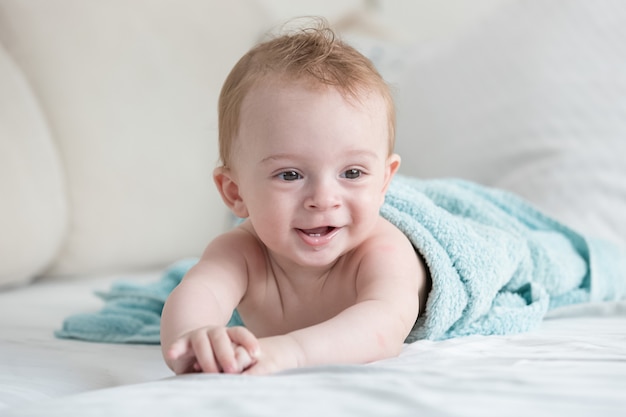 Portrait de bébé souriant dans une serviette bleue après la douche allongé sur le lit