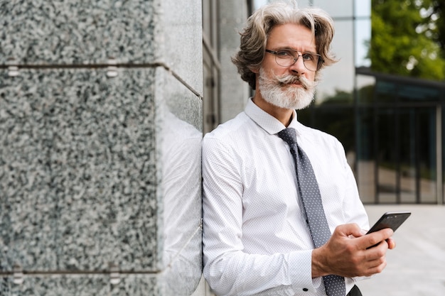 Portrait d'un bel homme d'affaires âgé à lunettes en tapant sur son téléphone portable tout en s'appuyant sur le mur à l'extérieur