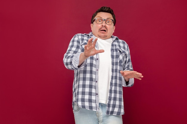 Portrait d'un bel homme d'affaires d'âge moyen choqué en chemise à carreaux décontractée et lunettes debout et regardant la caméra avec des yeux effrayés. tourné en studio intérieur, isolé sur fond rouge foncé.