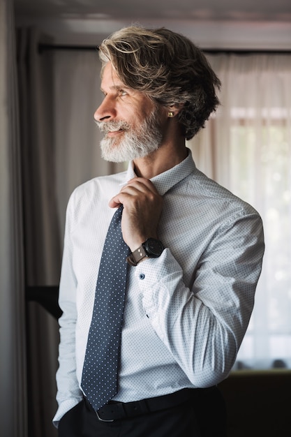 Portrait d'un bel homme d'affaires aux cheveux gris senior à l'intérieur à la maison vêtu de vêtements formels regardant la fenêtre.