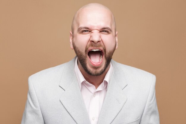 Portrait d'un bel homme d'affaires barbu chauve d'âge moyen en colère en costume gris clair classique debout et criant et regardant la caméra. tourné en studio intérieur, isolé sur fond marron clair.