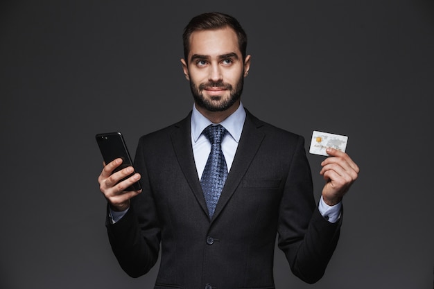 Portrait d'un bel homme d'affaires confiant portant costume isolé, à l'aide de téléphone mobile, montrant une carte de crédit en plastique