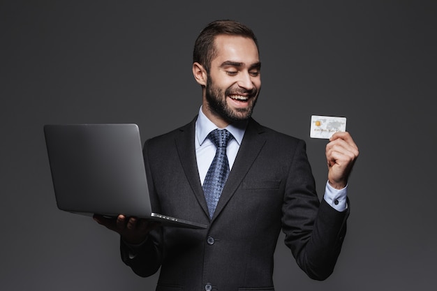 Portrait d'un bel homme d'affaires confiant portant costume isolé, tenant un ordinateur portable, montrant la carte de crédit