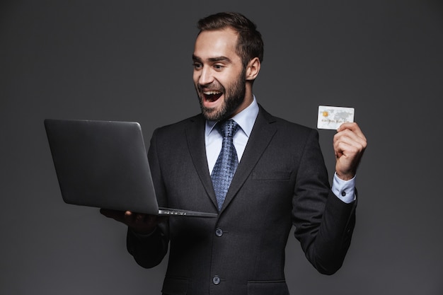 Portrait d'un bel homme d'affaires confiant portant costume isolé, tenant un ordinateur portable, montrant la carte de crédit