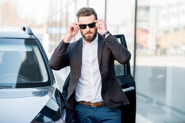 Portrait d'un bel homme d'affaires à lunettes de soleil debout près de la voiture à l'extérieur devant la façade du bâtiment moderne