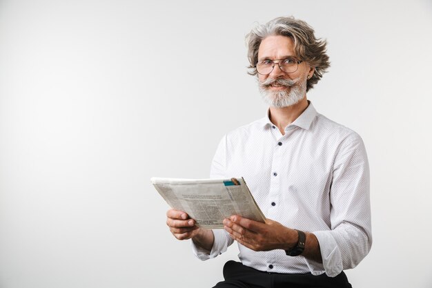 Portrait d'un bel homme d'affaires mature souriant vêtu de vêtements de cérémonie debout isolé sur un mur blanc, lisant le journal