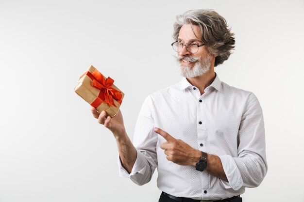 Portrait d'un bel homme d'affaires mature souriant vêtu de vêtements de cérémonie debout isolé sur un mur blanc, montrant la boîte actuelle, pointant le doigt