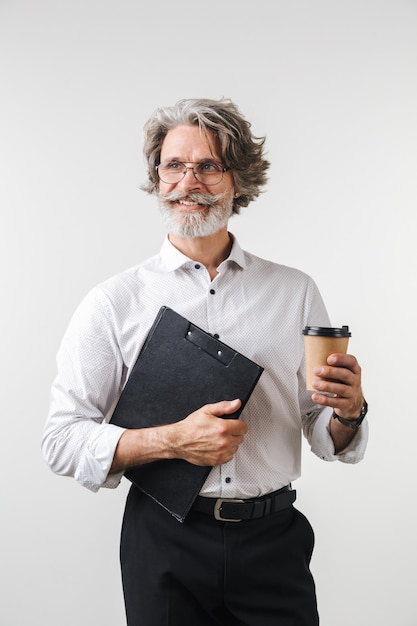 Portrait d'un bel homme d'affaires mature souriant vêtu de vêtements de cérémonie debout isolé sur un mur blanc, tenant un bloc-notes, buvant du café à emporter