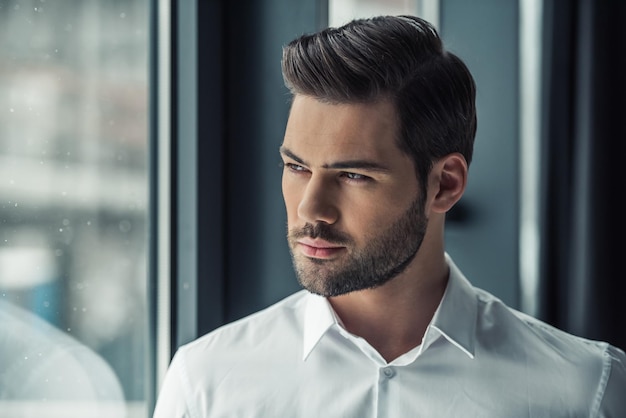 Portrait d'un bel homme d'affaires regardant par la fenêtre et pensant en se tenant au bureau