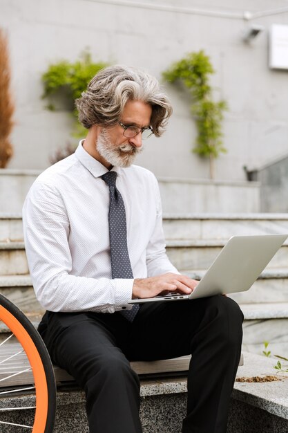 Portrait d'un bel homme d'affaires sérieux à lunettes en tapant sur un ordinateur portable assis sur un banc avec un vélo à l'extérieur