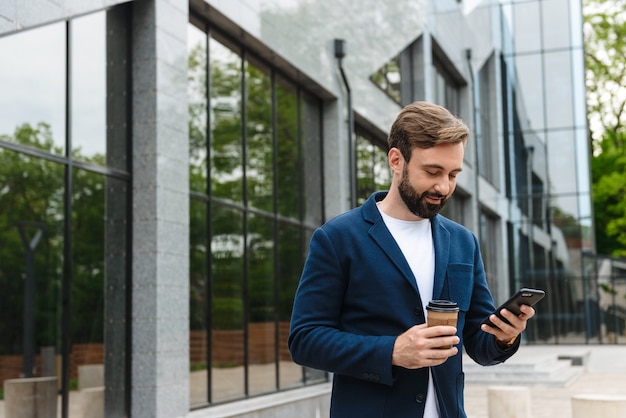 Portrait d'un bel homme d'affaires en veste tenant un téléphone portable tout en se tenant à l'extérieur près du bâtiment avec du café à emporter