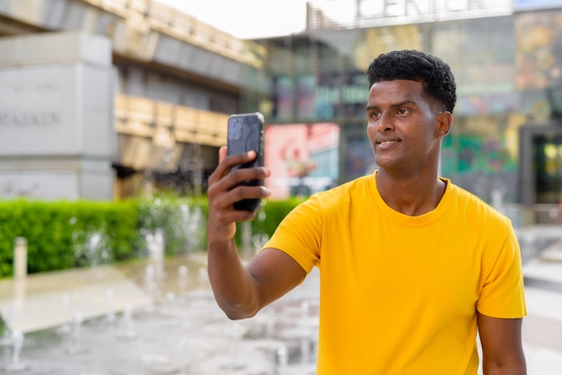 Portrait d'un bel homme africain noir portant un t-shirt jaune à l'extérieur de la ville pendant l'été à côté de la fontaine tout en utilisant un téléphone portable