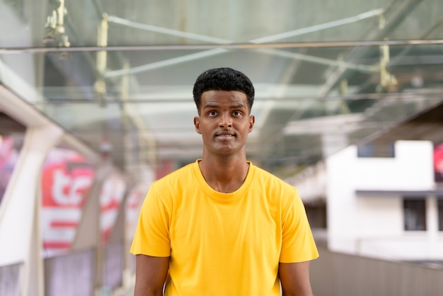 Portrait d'un bel homme africain noir portant un t-shirt jaune à l'extérieur de la ville pendant l'été tout en regardant devant la passerelle