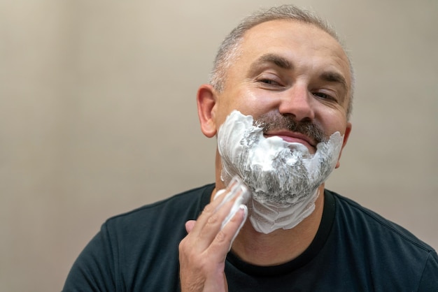 Portrait d'un bel homme d'âge moyen barbu aux cheveux blancs appliquant de la mousse à raser pour couper sa barbe