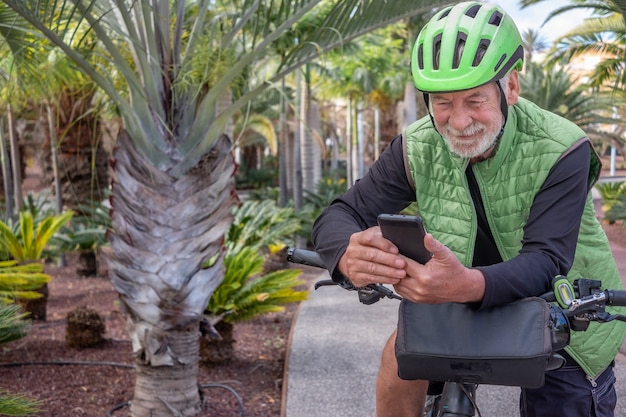 Portrait d'un bel homme âgé utilisant un téléphone intelligent lors d'une balade à vélo dans un lieu tropical extérieur. Retraité aux cheveux blancs profitant de son temps libre