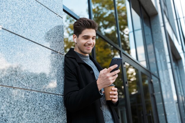 Portrait d'un bel homme des années 30 portant des écouteurs tenant un smartphone et du café à emporter en se tenant devant un bâtiment en verre