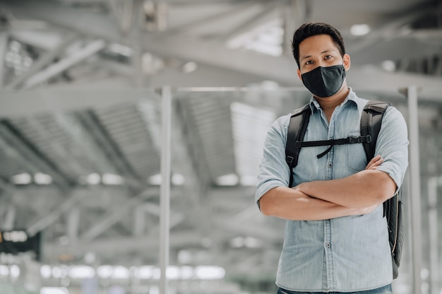 Portrait d'un bel homme asiatique avec un masque facial pour se protéger du coronavirus en chemise bleue debout avec un sac à dos les bras croisés regardant la caméra