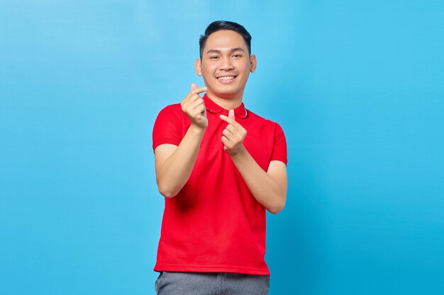 Portrait d'un bel homme asiatique souriant en chemise rouge montrant le cœur avec les doigts croisés, exprimant la joie et la positivité isolé sur fond bleu