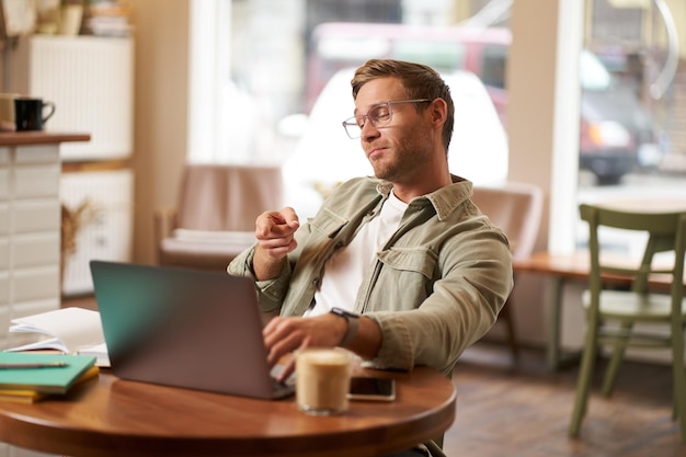 Portrait d'un bel homme aux lunettes assis dans un café pointe le doigt sur l'écran de l'ordinateur portable les discussions vidéo se connectent