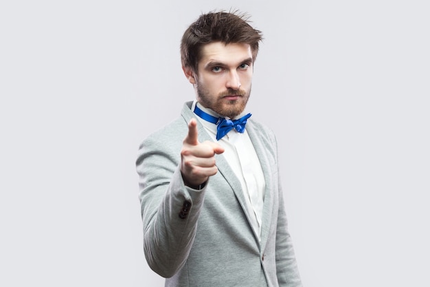 Portrait d'un bel homme barbu autoritaire sérieux en costume gris décontracté et noeud papillon bleu debout, regardant la caméra et se réchauffant. tourné en studio intérieur, isolé sur fond gris clair.