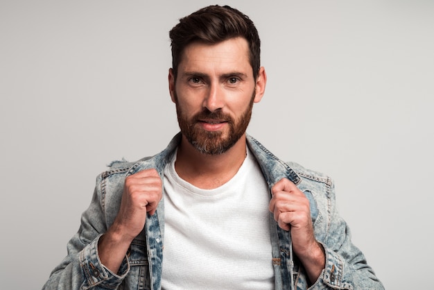 Portrait d'un bel homme barbu dans des vêtements décontractés regardant la caméra et souriant tout en corrigeant sa veste et en posant. Studio tourné sur fond blanc