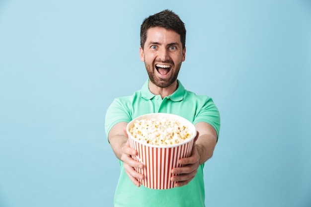 Portrait d'un bel homme barbu portant des vêtements décontractés, isolé sur un mur bleu, mangeant du pop-corn en regardant un film