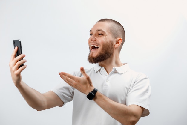 Portrait d'un bel homme barbu positif dans un t-shirt blanc parlant sur appel vidéo à l'aide d'un smartphone