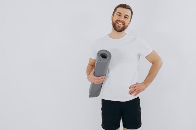 Portrait d'un bel homme barbu en vêtements de sport tenant un tapis de yoga sur l'espace de copie de fond blanc