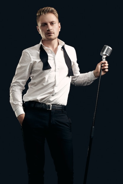 Portrait de bel homme blond chanteur en smoking élégant et noeud papillon posant avec microphone vintage sur mur noir