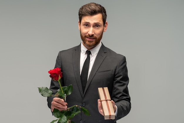 Portrait d'un bel homme brune barbu en costume formel debout tenant une fleur de rose rouge et une boîte avec le présent tourné en studio intérieur isolé sur fond gris