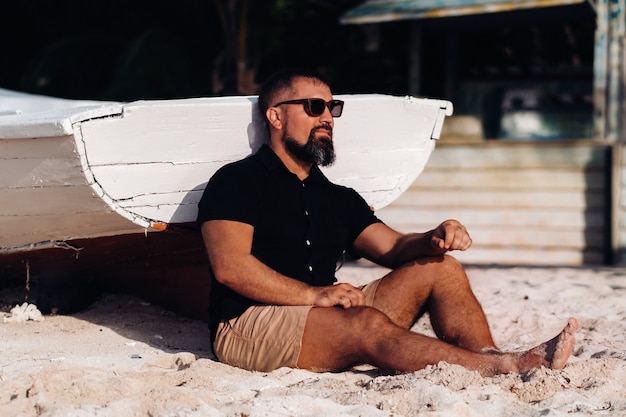 Portrait D'un Bel Homme Brutal, Appréciant Et Relaxant, Assis Sur La Plage De L'île Maurice. L'homme Sur La Plage Du Morne Dans Les Vêtements Noirs Et Les Lunettes. Plage Tropicale.