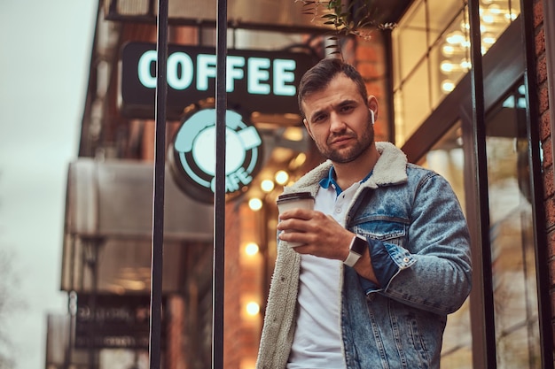 Portrait d'un bel homme élégant portant une veste en jean avec des écouteurs sans fil tenant du café à emporter à l'extérieur du café.