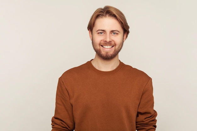 Portrait d'un bel homme heureux avec des cheveux et une barbe soignés portant un sweat-shirt debout, regardant la caméra