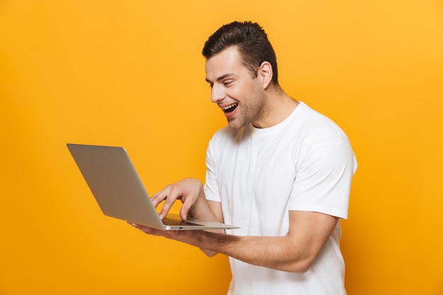 Portrait d'un bel homme heureux portant un t-shirt debout isolé sur un mur jaune, utilisant un ordinateur portable, célébrant