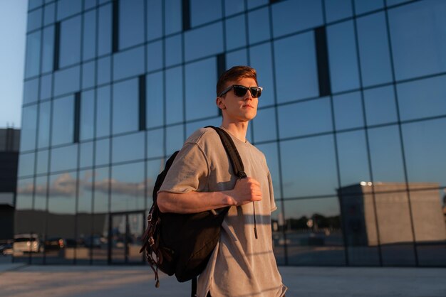 Portrait d'un bel homme hipster touristique à lunettes de soleil avec sac à main mec debout près de l'immeuble au centre-ville Calme moment de détente concept de vacances d'été