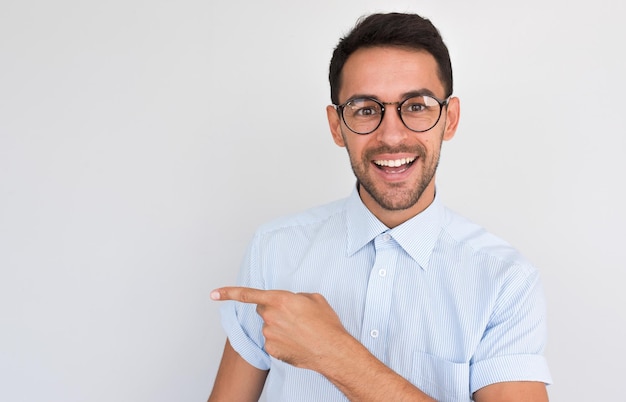 Portrait d'un bel homme mal rasé heureux souriant avec un sourire à pleines dents pointe de côté en regardant vers la caméra montre l'espace de copie pour votre contenu publicitaire contre le mur blanc du studio en chemise bleue décontractée