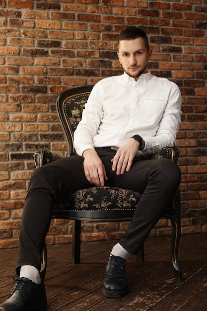 Portrait bel homme à la mode dans une chemise blanche est assis sur une chaise dans un loft de studio photo. Jeune homme d'affaires