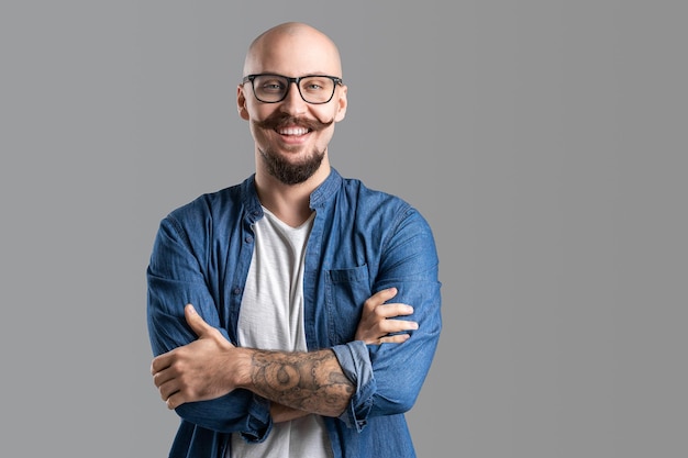 Portrait d'un bel homme à la mode heureux en chemise jeans croisant les mains et regardant la caméra en souriant tout en posant sur fond de studio gris