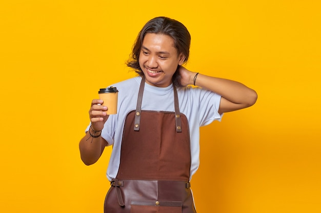 Portrait d'un bel homme portant un tablier regardant une tasse de café se tenant isolé sur fond jaune