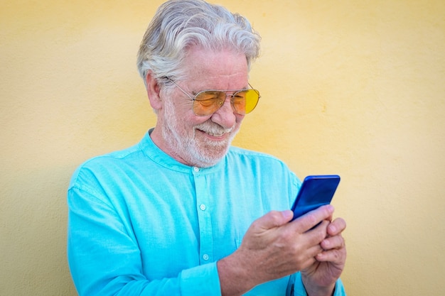 Portrait d'un bel homme senior en plein air, debout contre un mur jaune à l'aide d'un téléphone portable