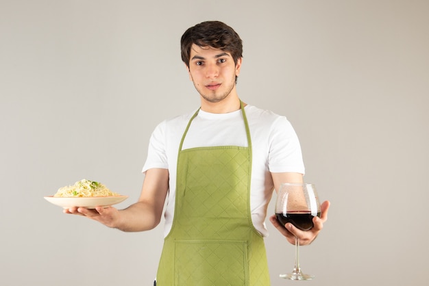 Portrait d'un bel homme en tablier tenant une assiette avec des nouilles et un verre de vin.