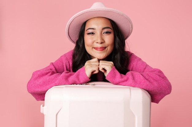 Portrait de belle belle fille asiatique portant un chapeau souriant et tenant une valise de voyage isolée sur rose